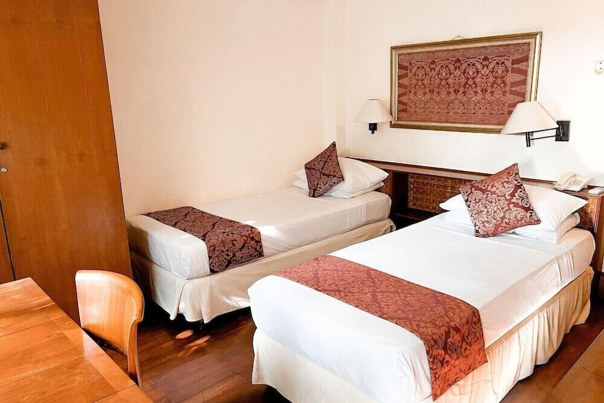 Una habitación de hotel con dos camas, un escritorio de madera, una silla, una lámpara de pared y una obra de arte enmarcada sobre las camas.