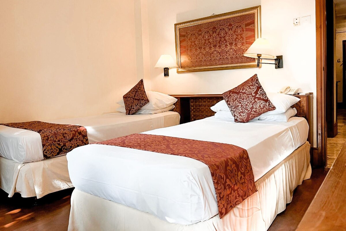 A hotel room with two beds, each with a patterned bedspread and pillows, a wall-mounted lamp, and a framed artwork above the beds.