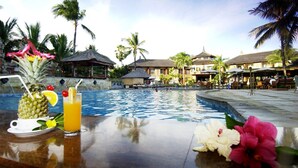 Ambiente de resort tropical à beira da piscina, com piscina, palmeiras e área de refeições com guarda-sóis.