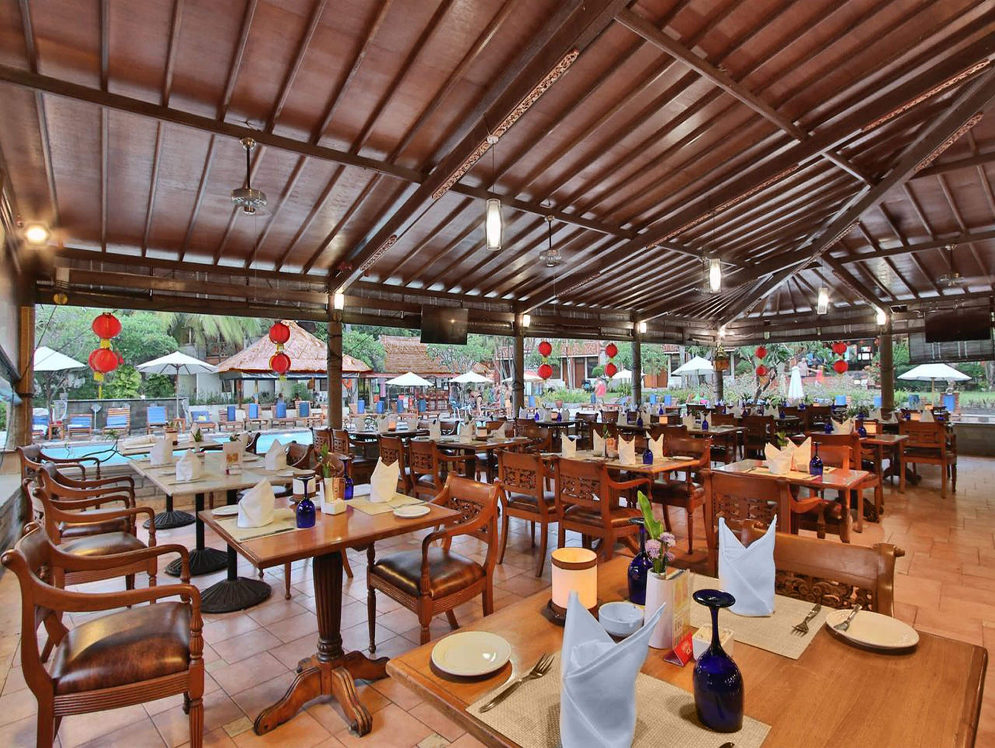 Un restaurante al aire libre con mesas y sillas de madera, sombrillas y una piscina al fondo.