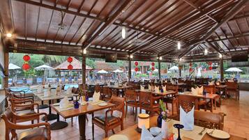 Un restaurante al aire libre con mesas y sillas de madera, sombrillas y una piscina al fondo.
