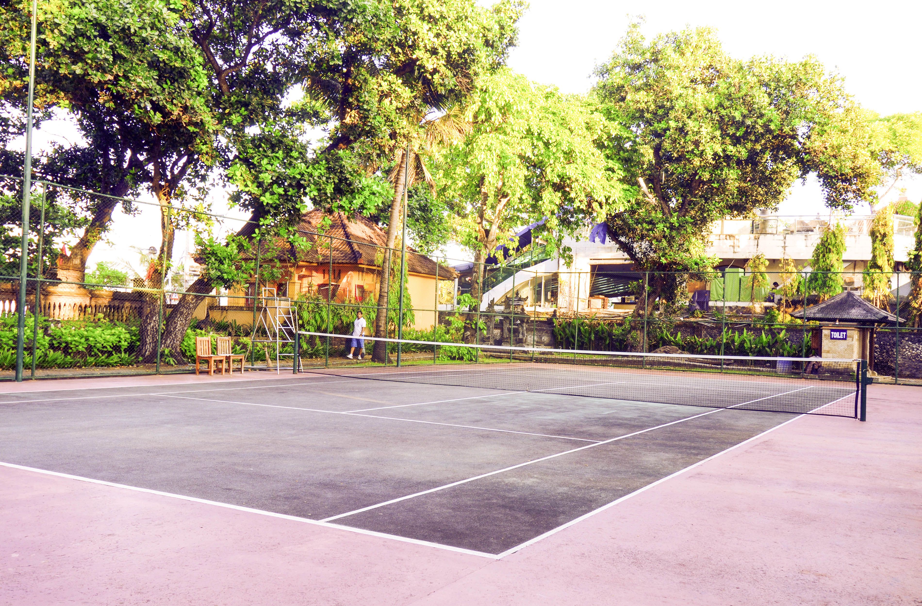 An outdoor tennis court with a net, surrounded by trees and a small pavilion.