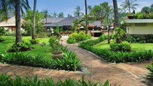 Um resort tropical com um calçadão pavimentado, vegetação exuberante, palmeiras e edifícios de estilo tradicional.
