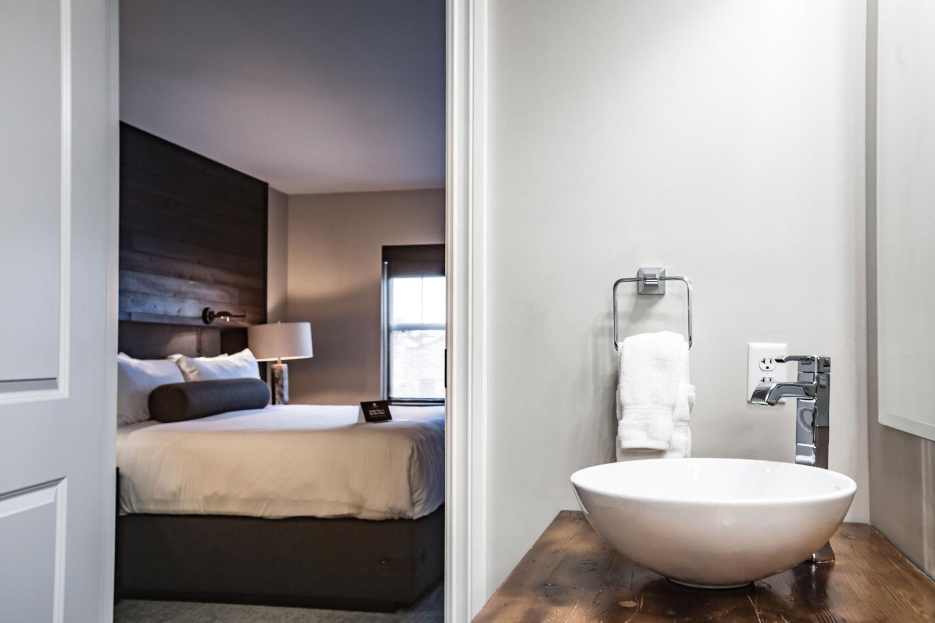 A modern hotel room with a large bed, bedside lamp, and a bathroom with a large sink and towel rack.