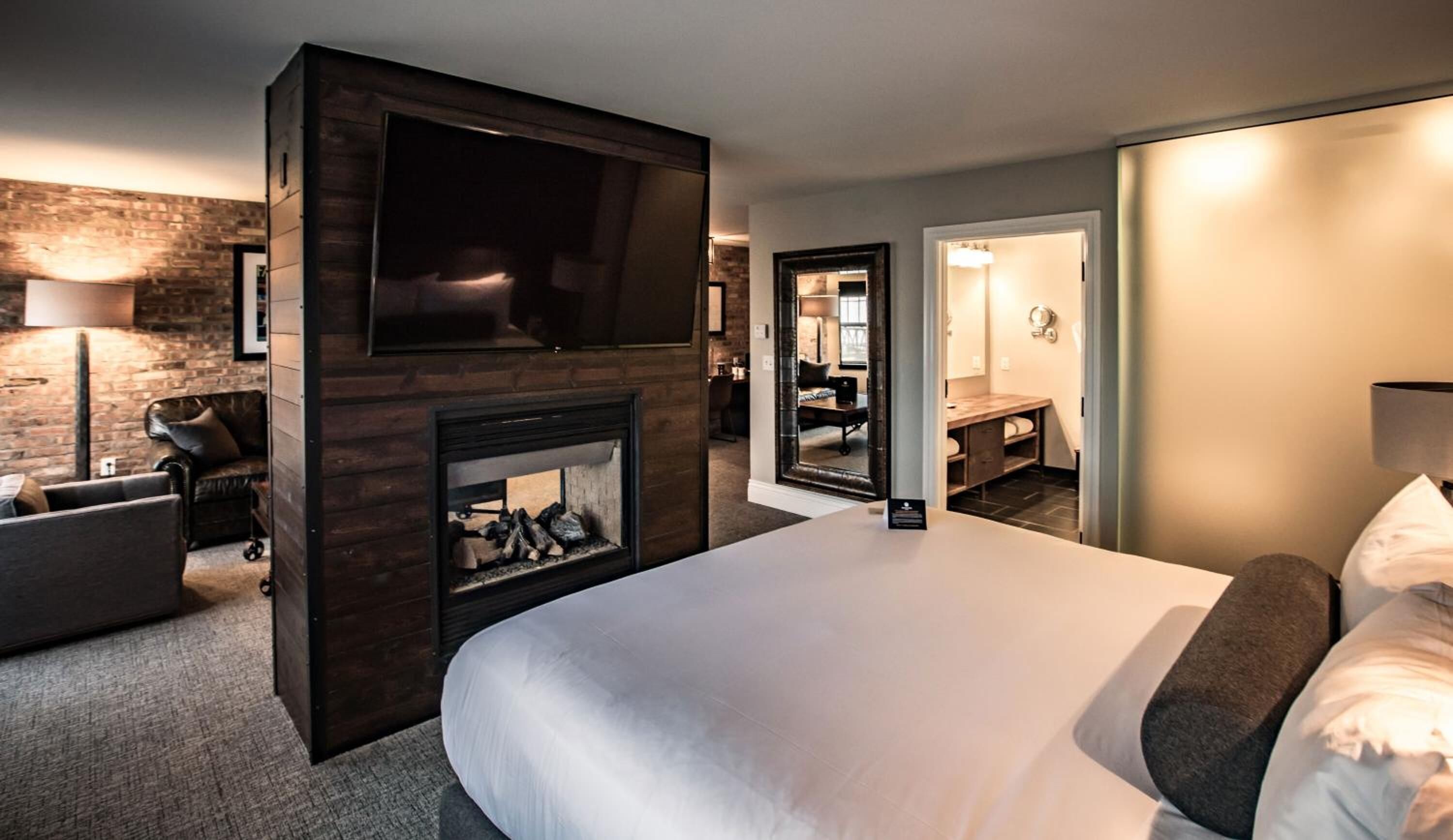 A hotel room with a large bed, a fireplace, a TV, a desk, and a bathroom visible through an open door.