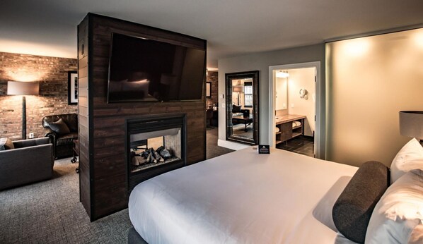 Habitación de hotel con una cama grande, chimenea, televisor, escritorio y baño visible a través de una puerta abierta.