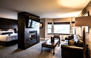 A modern hotel room with a bed, a fireplace, a leather sofa, and a large window.