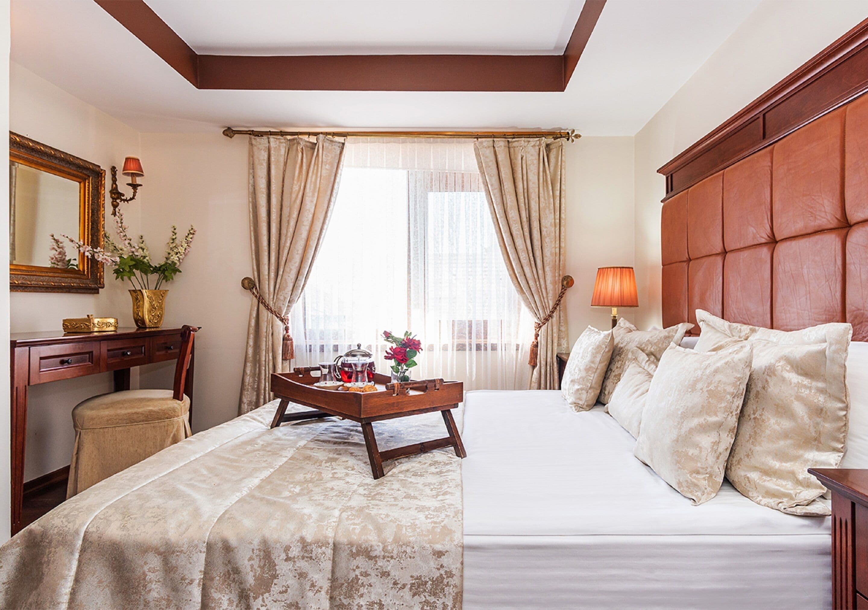 A hotel room with a large bed, a wooden desk, a chair, a bedside table with a lamp, and a window with curtains.