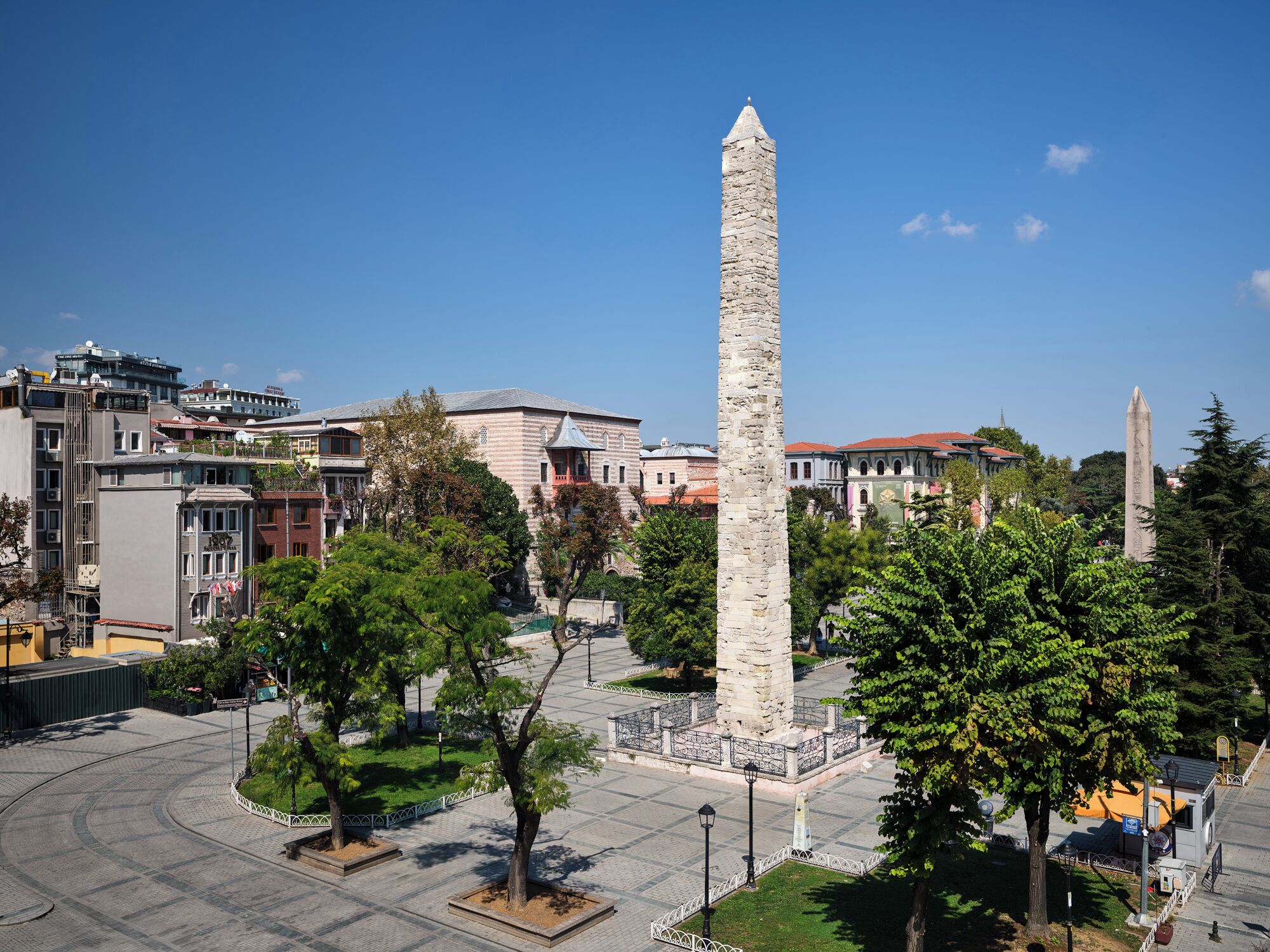 Un alto obelisco de piedra en una plaza rodeada de árboles y edificios.