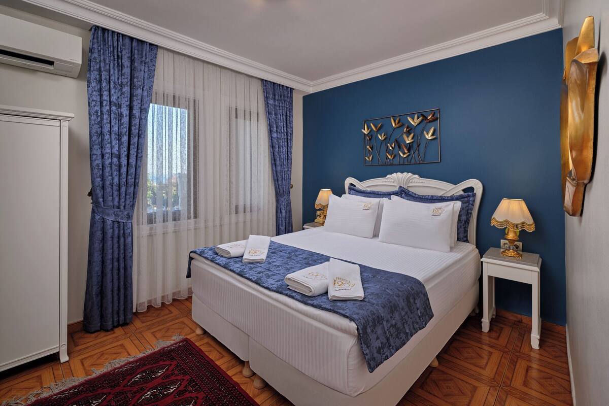 A hotel room with a large bed, two bedside tables, a chandelier, and a view of the outdoors through sheer curtains.