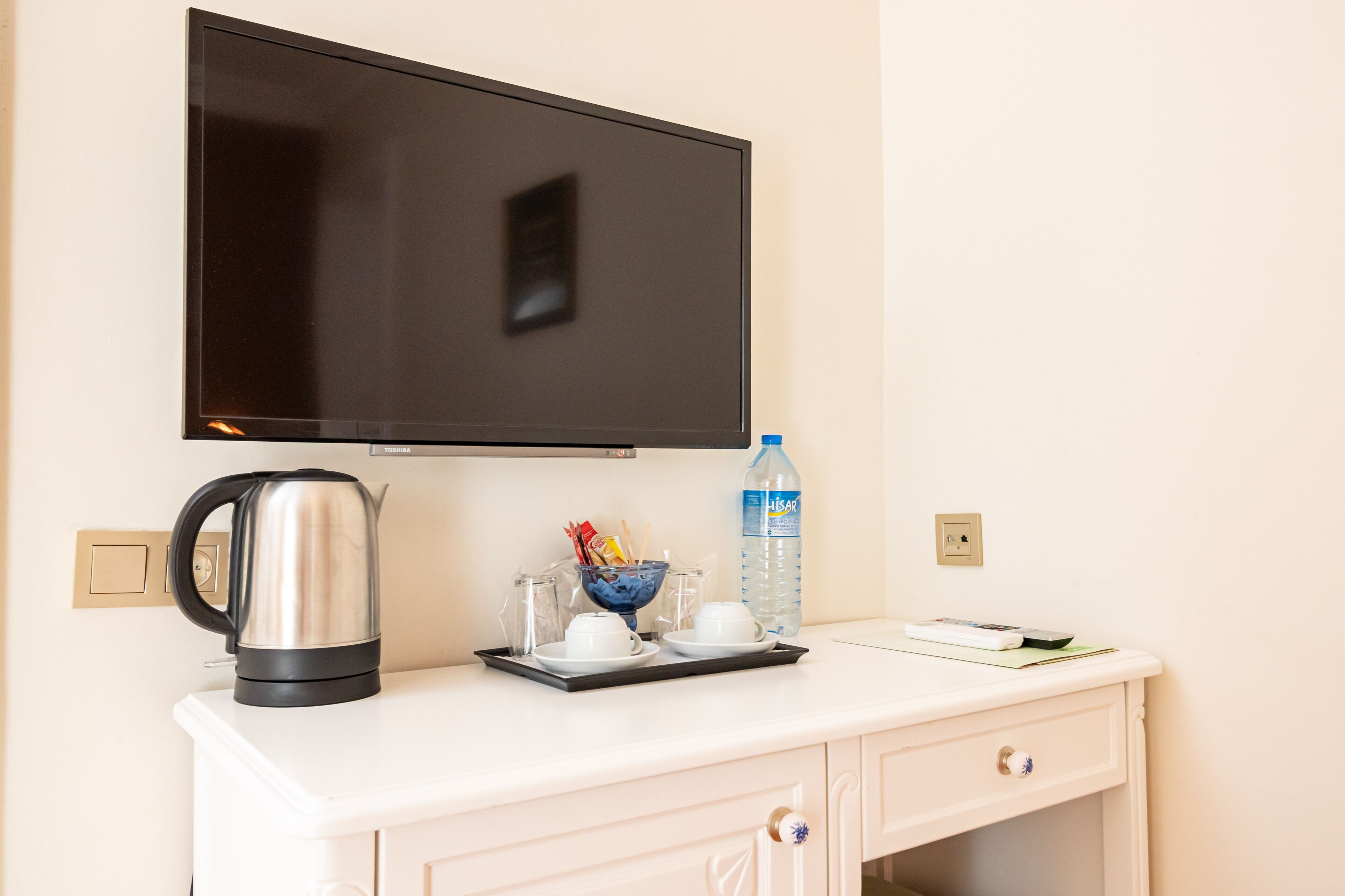 Une chambre d’hôtel équipée d’un téléviseur à écran plat fixé au mur, d’un bureau blanc avec une bouilloire, des tasses, une bouteille d’eau et un livre.
