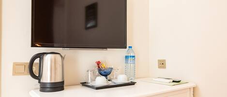 Une chambre d’hôtel équipée d’un téléviseur à écran plat fixé au mur, d’un bureau blanc avec une bouilloire, des tasses, une bouteille d’eau et un livre.