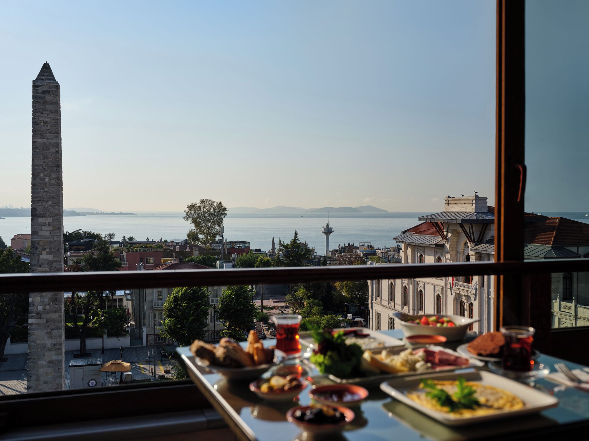 A table with various dishes and drinks, overlooking a cityscape with a prominent tower and a body of water.