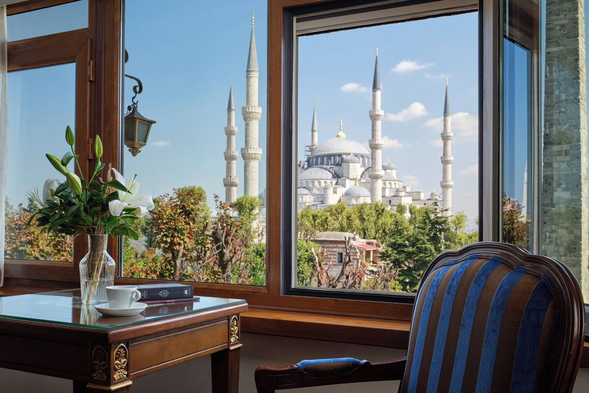 A room with a view of a mosque, a wooden desk with a vase of flowers, and a striped armchair.