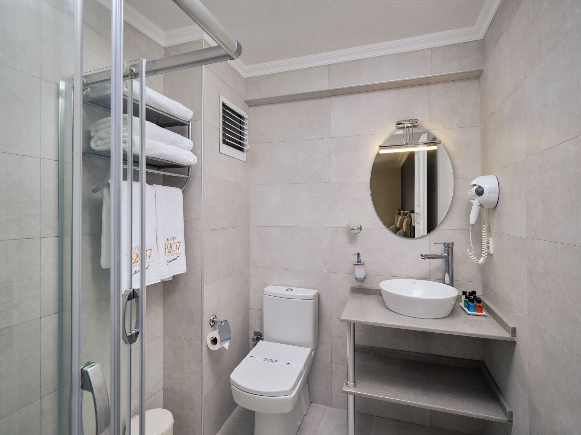 A modern bathroom with a glass shower enclosure, a toilet, a sink with a mirror, and a hairdryer mounted on the wall.