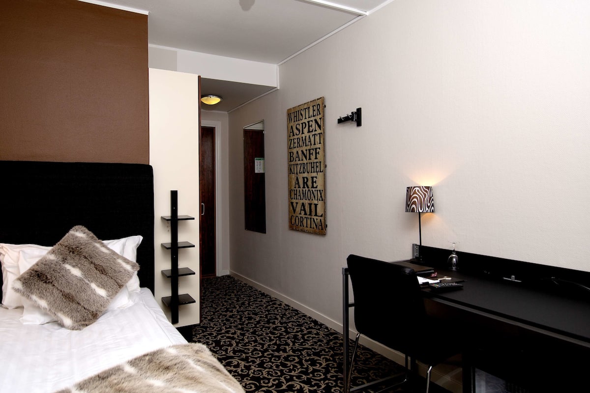 A hotel room with a bed, a desk, a lamp, and a wall with a sign listing names.