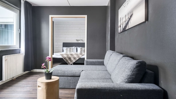 A modern living room with a grey sectional sofa, a wooden side table with a potted plant, and a large window with curtains. - Sure Hotel by Best Western Algen (Östersund)