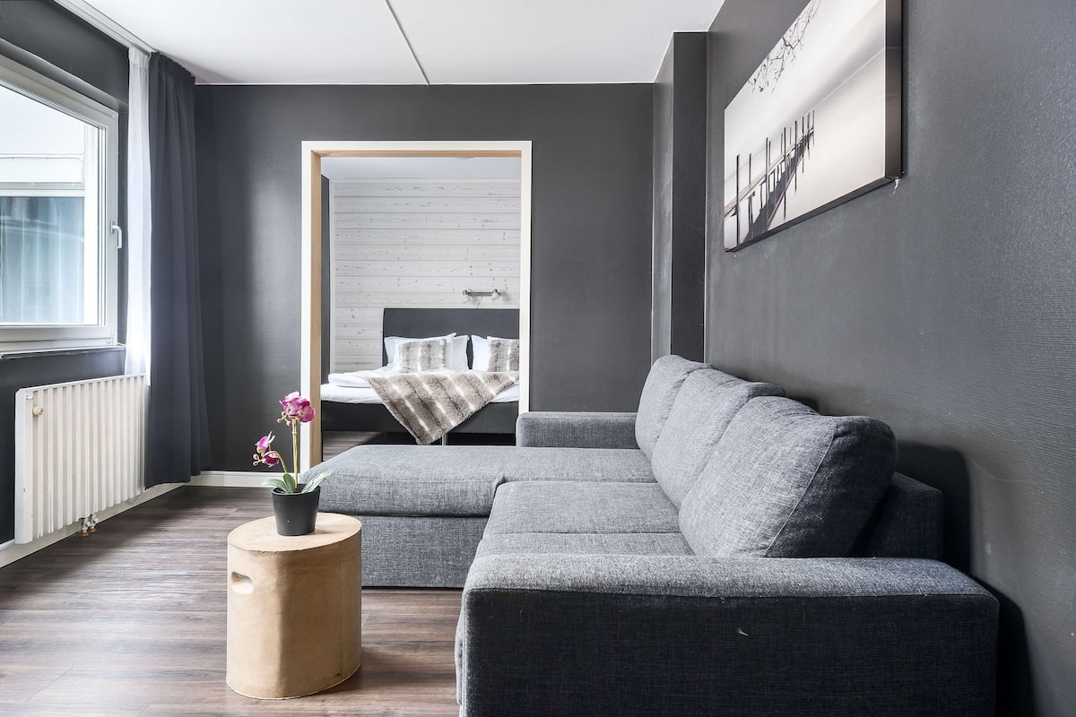 A modern living room with a grey sectional sofa, a wooden side table with a potted plant, and a large window with curtains.