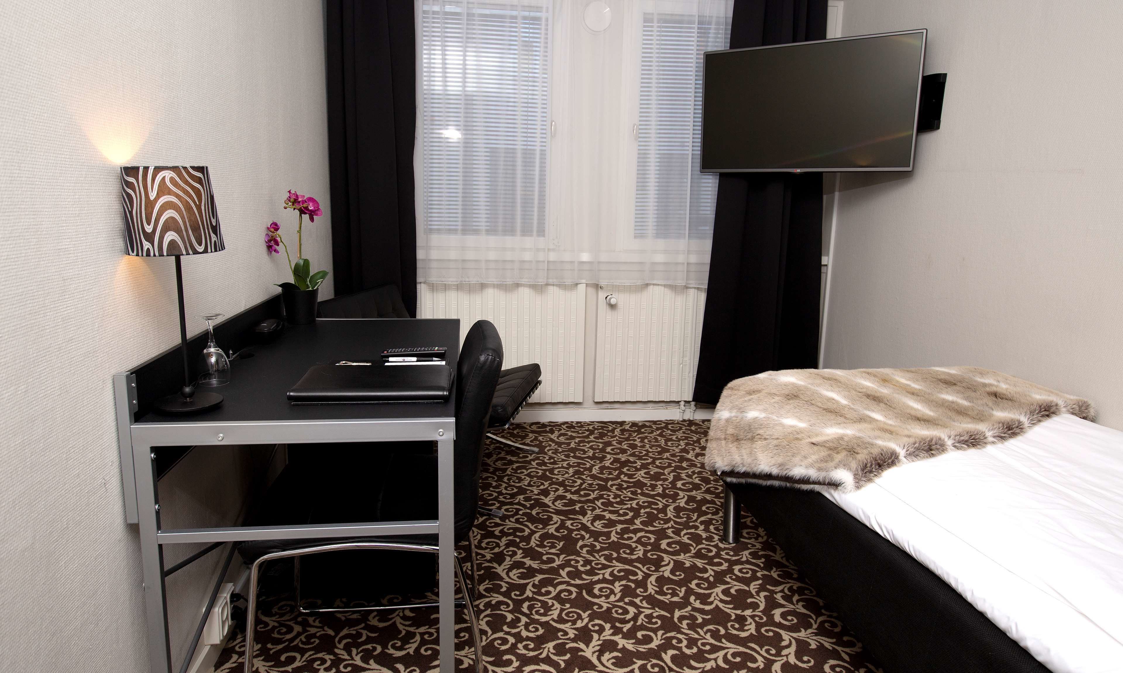 Ein Hotelzimmer mit Bett, Schreibtisch mit Stuhl, flachbildschirmfernseher an der Wand, kleiner Tisch mit Lampe und Zimmerpflanze.