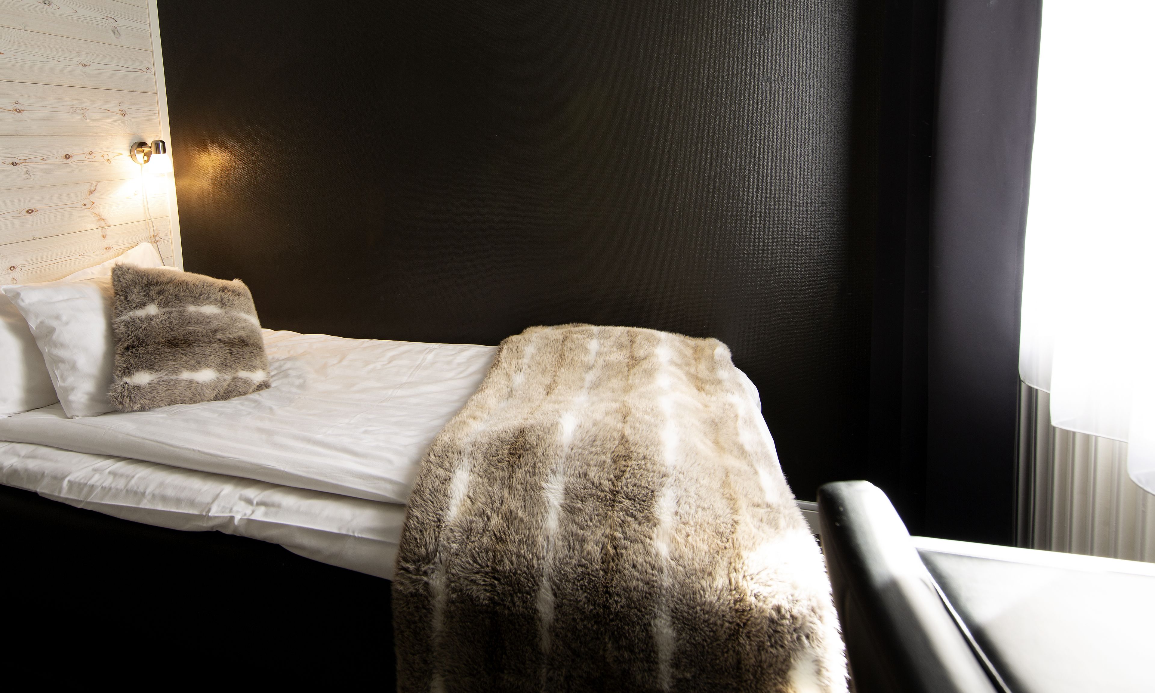A single bed with white bedding and a fur throw, a nightstand with a lamp, and a wall with a wooden panel design.