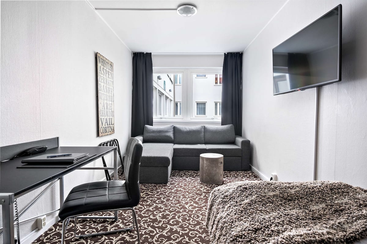 A modern hotel room with a desk, chair, sofa, and TV.