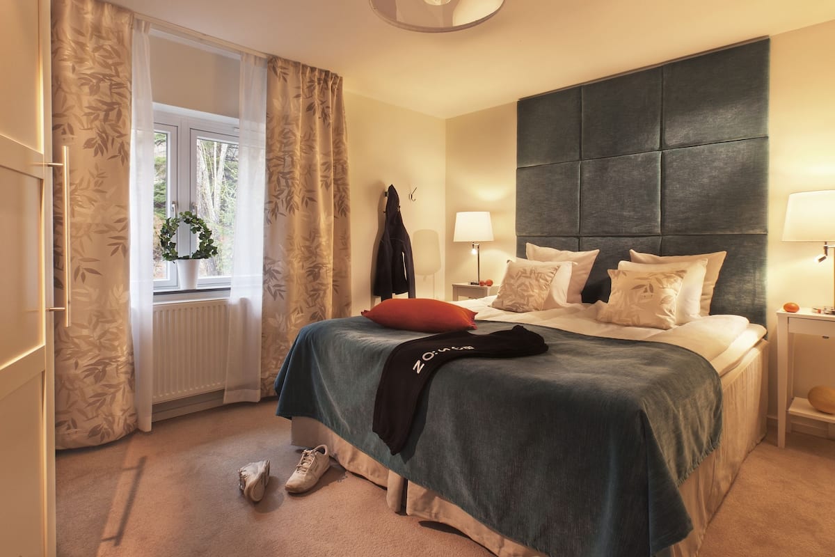 A hotel room with a large bed, two bedside tables with lamps, a window with curtains, a radiator, and a plant.
