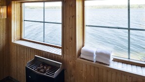 A wooden cabin with two windows overlooking a body of water. There is a wooden bench with towels on it, and a black trash bin with potatoes visible. - Bosön Hotell & Konferens (Lidingo)