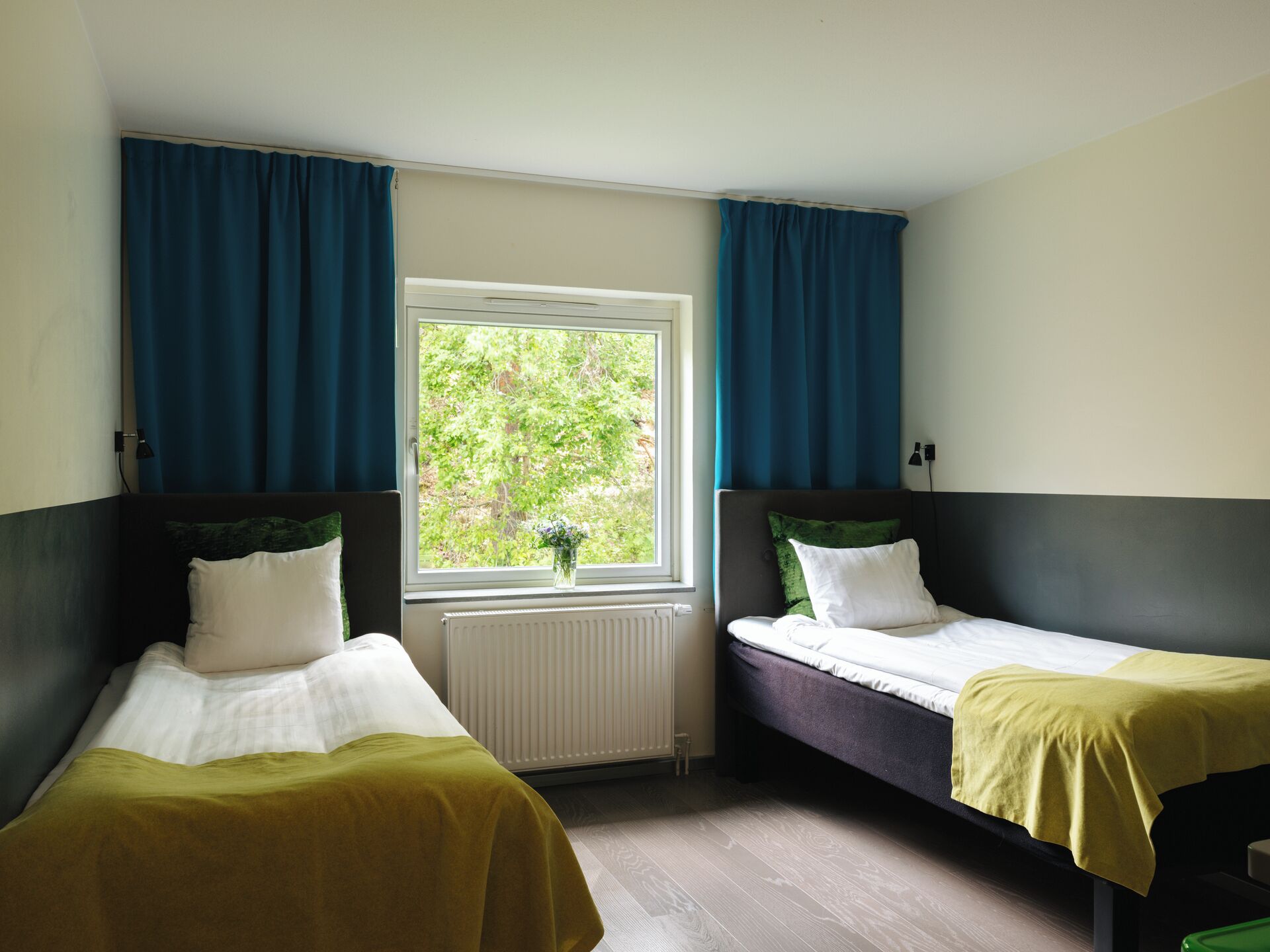Quarto com duas camas de solteiro, janela com cortinas, radiador e vista para o verde lá fora.