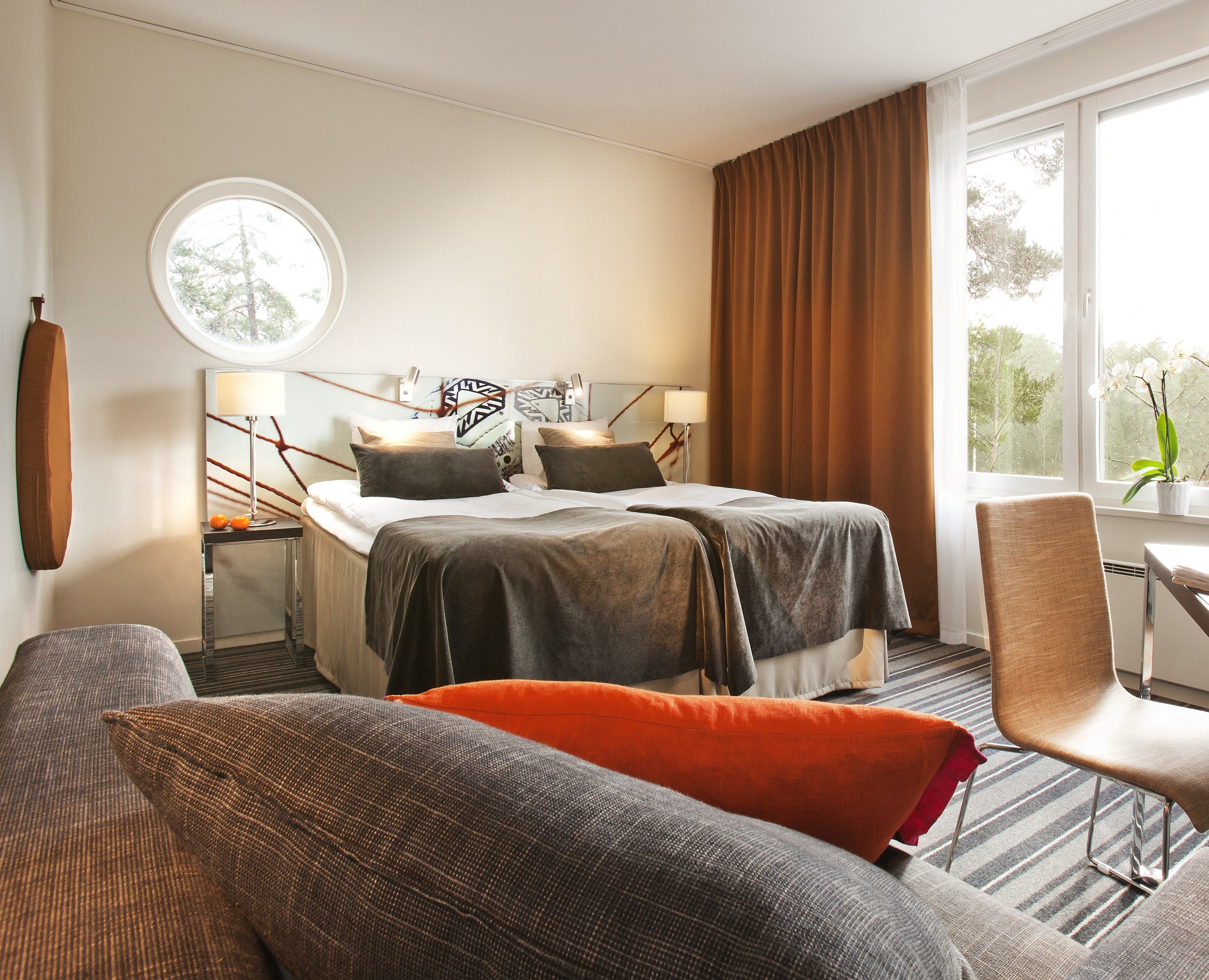 Une chambre d’hôtel avec un lit, un bureau, une chaise, une fenêtre ronde et une vue sur les arbres.