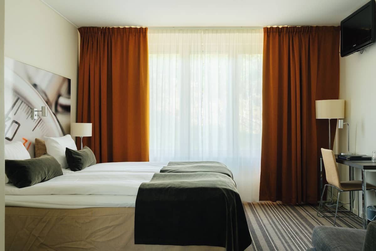 A hotel room with two beds, a desk, a chair, a TV, and a large window with curtains.