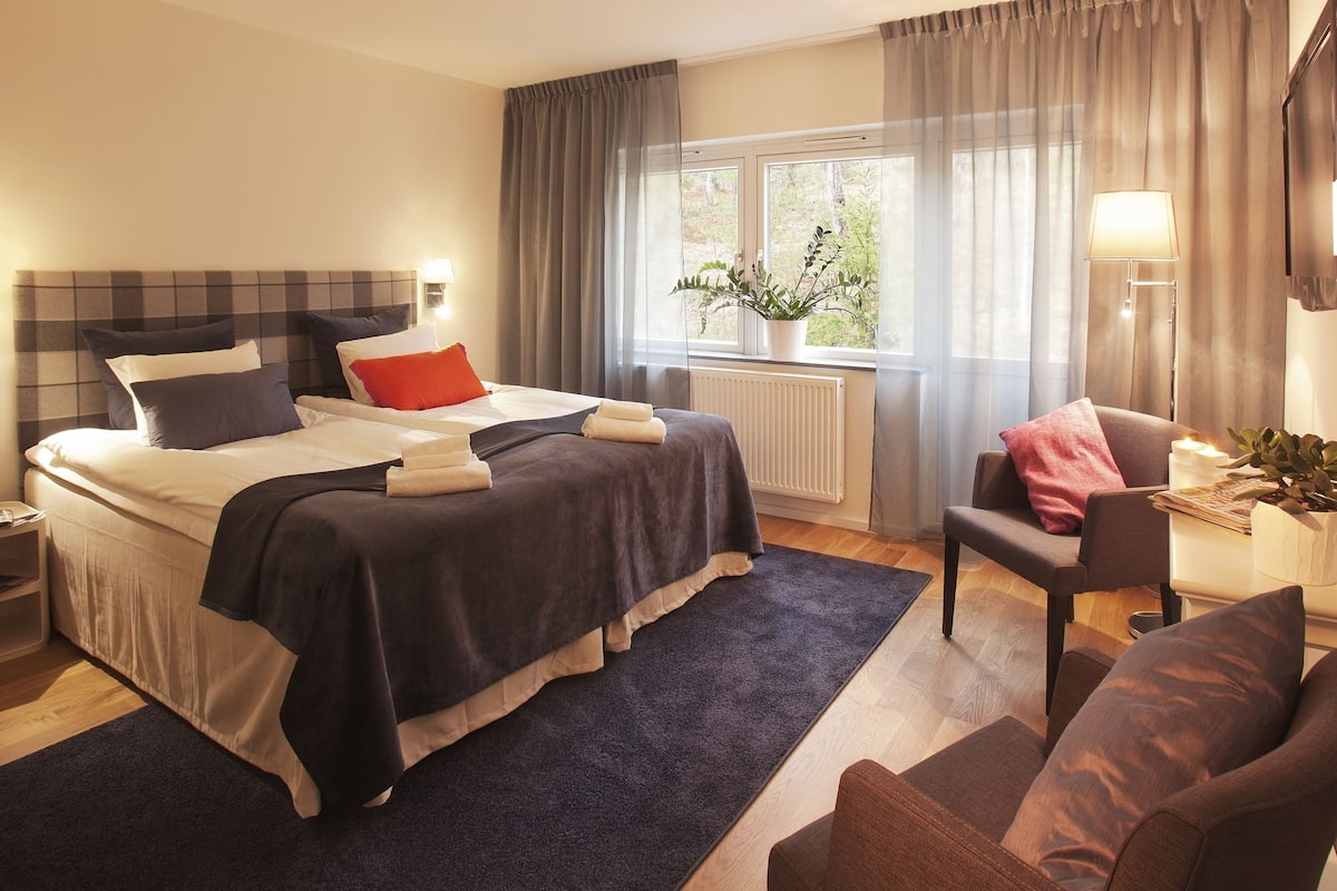 A hotel room with a bed, a chair, a window with a view of greenery, a radiator, and a lamp.