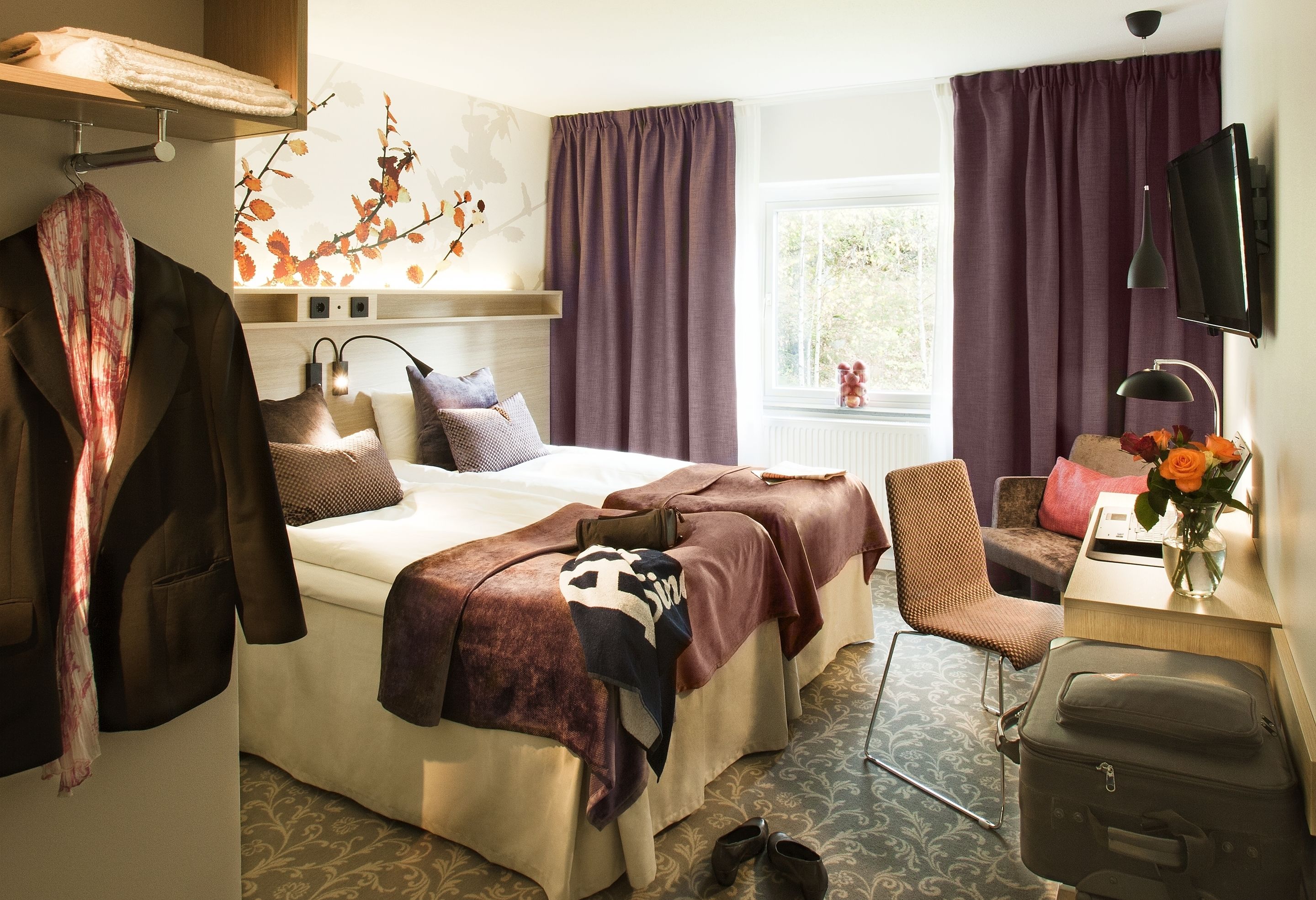 A hotel room with a bed, a desk with a computer, a chair, a suitcase, and a TV on the wall.