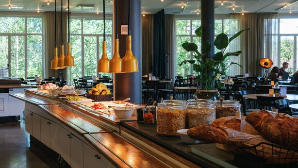 A buffet area with various food items, including fruits, bread, and cereal, in a well-lit room with large windows and hanging pendant lights. - Bosön Hotell & Konferens (Lidingo)