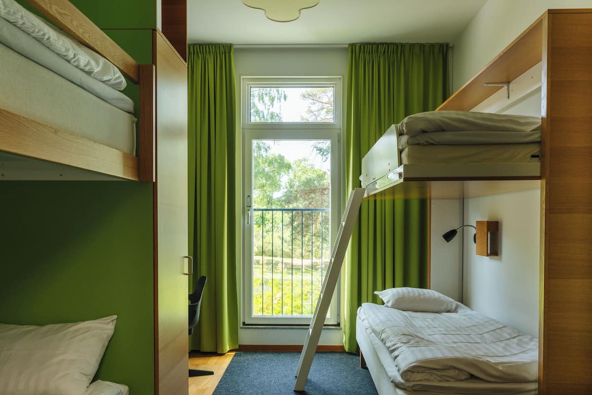 A dormitory room with bunk beds, a window with curtains, and a view of greenery outside.