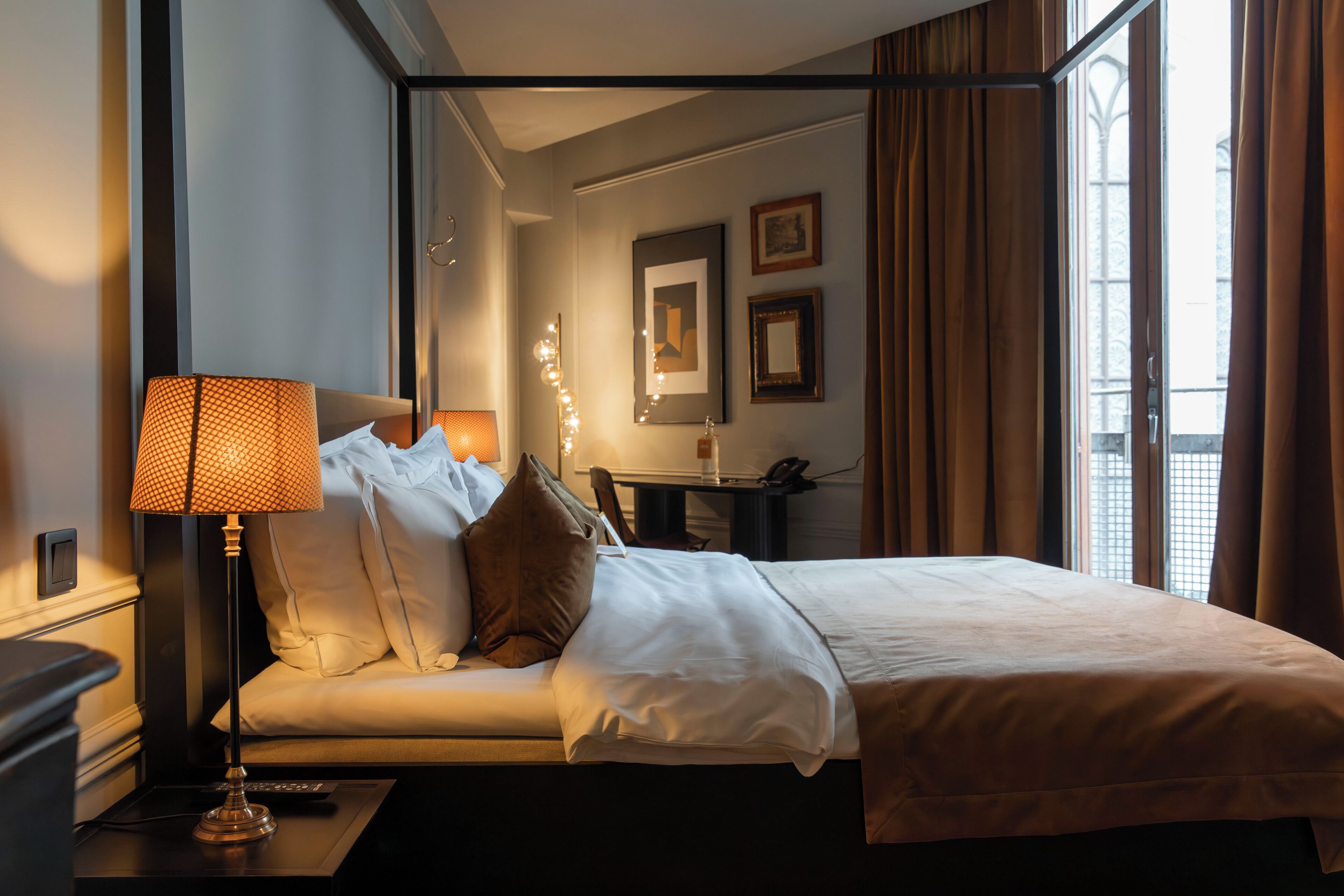 Habitación de hotel con cama, mesita de noche con lámpara, escritorio con silla y ventana con cortinas.