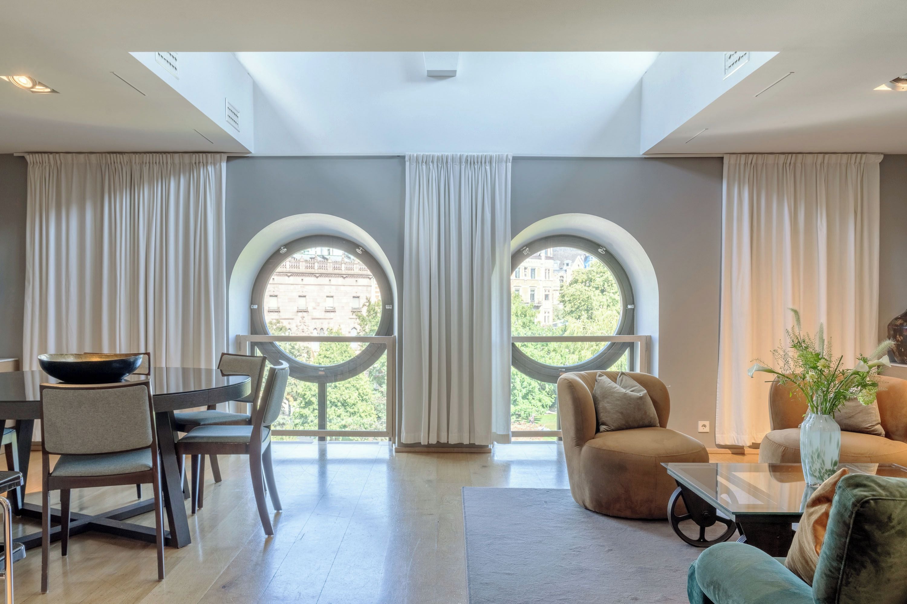 A modern living room with a dining area, featuring a large skylight, two arched windows, and a view of buildings.