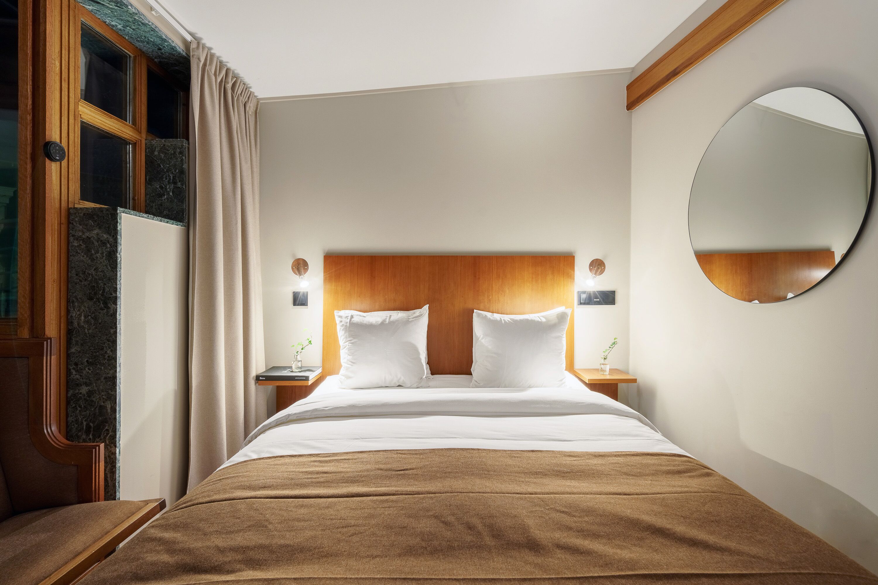 A hotel room with a wooden headboard, a large round mirror, a bed with white linens, and a brown throw blanket.