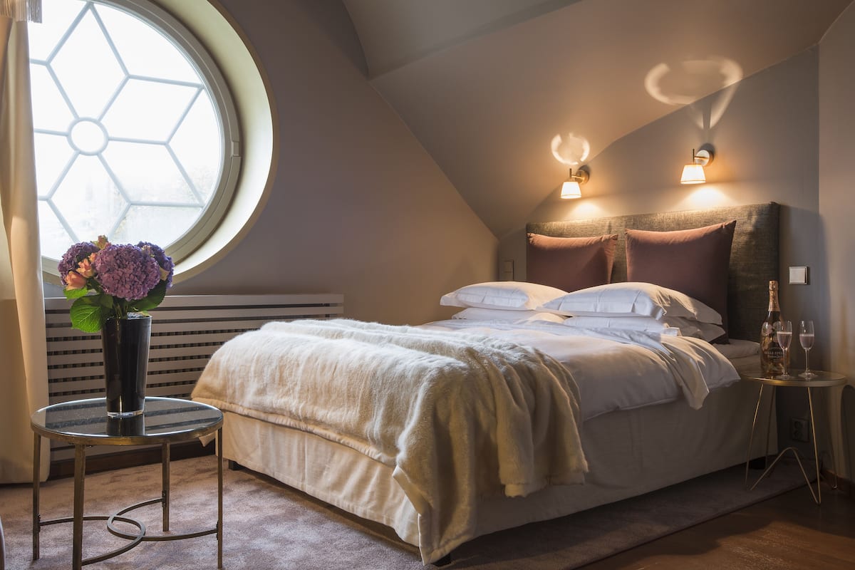 A bedroom with a bed, a round window, a vase with flowers, and wall-mounted lights.