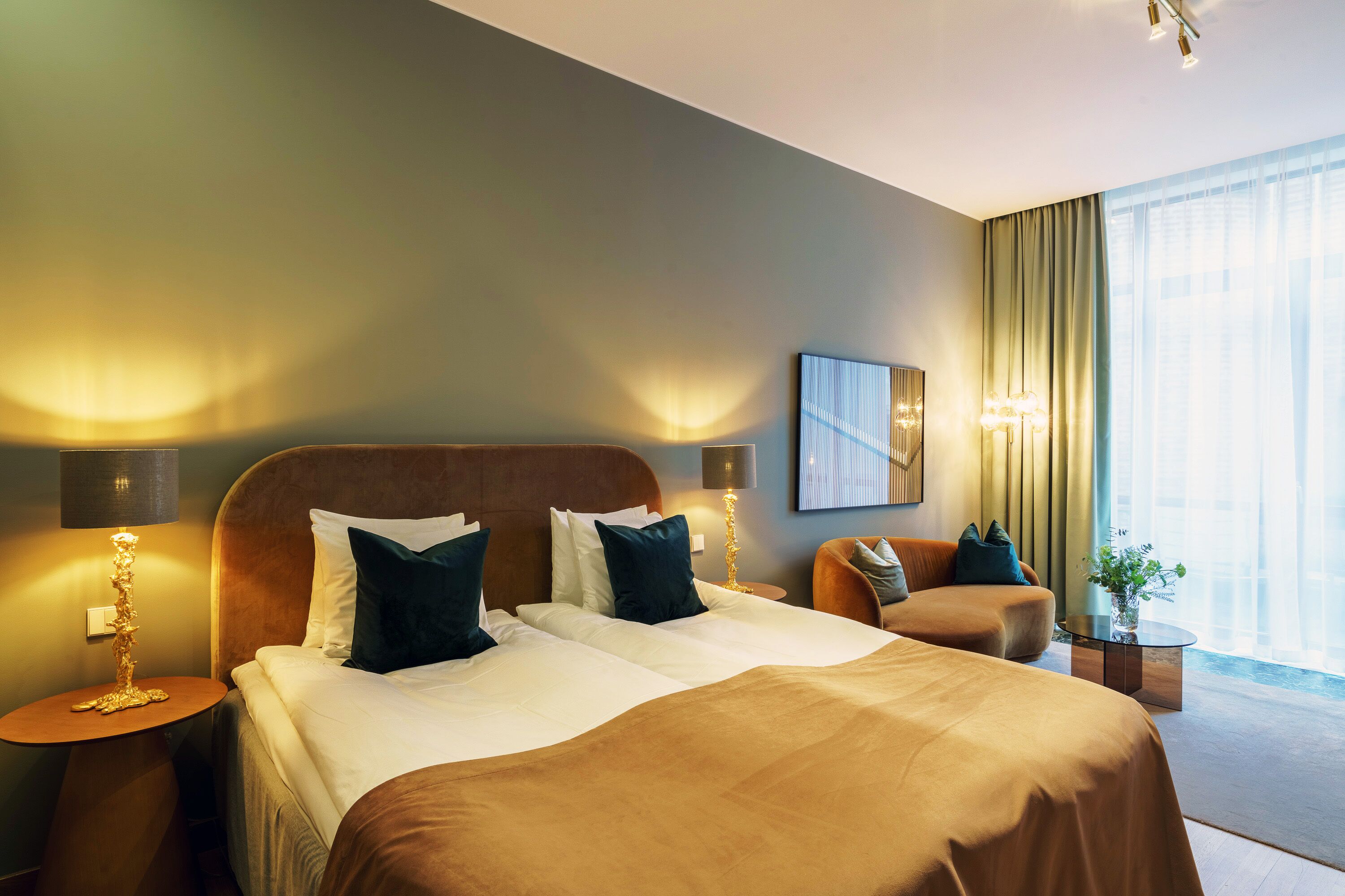 A modern hotel room with a large bed, two bedside tables, a seating area with a sofa, and a large window with curtains.