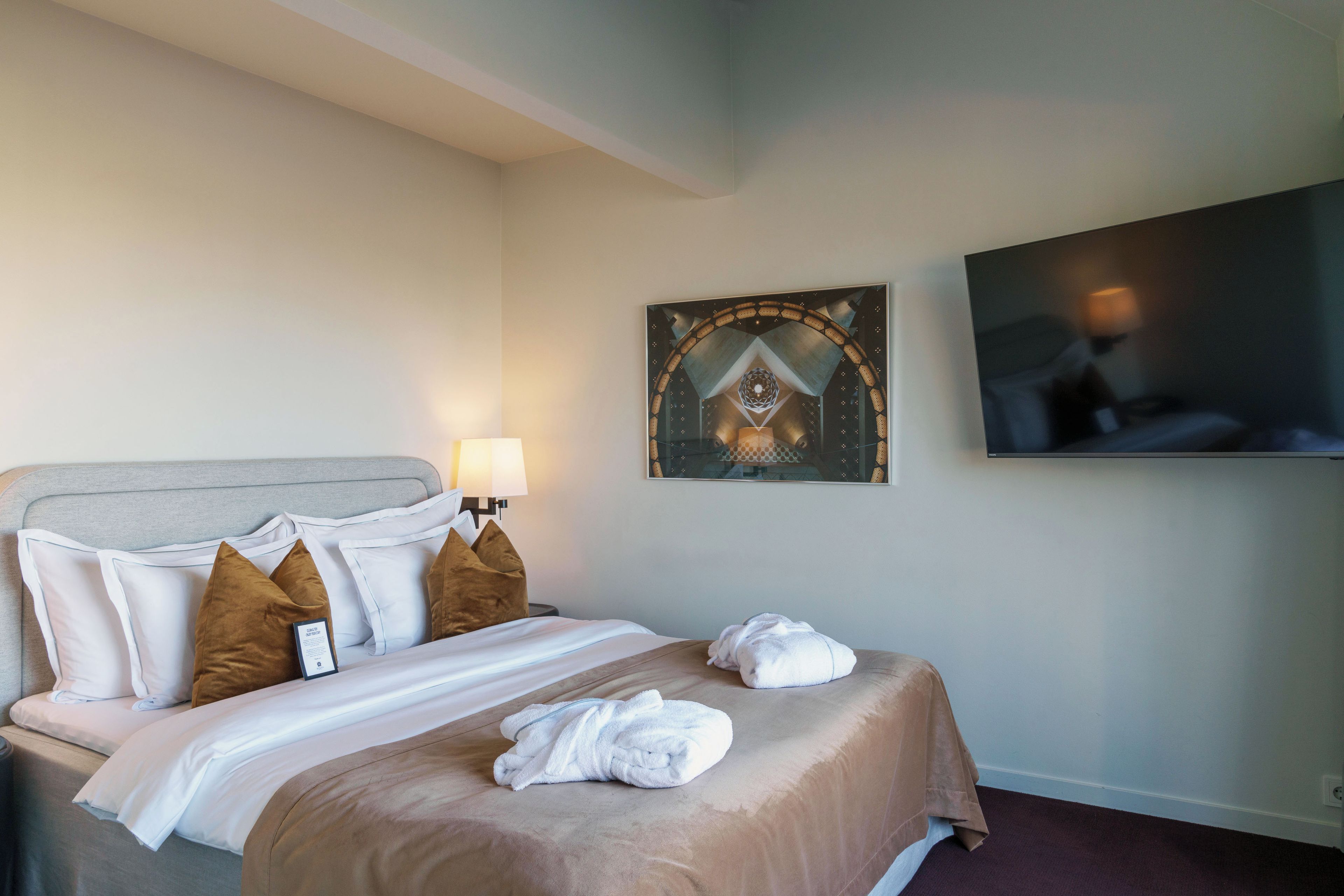 A hotel room with a bed, pillows, a TV, and a painting on the wall.