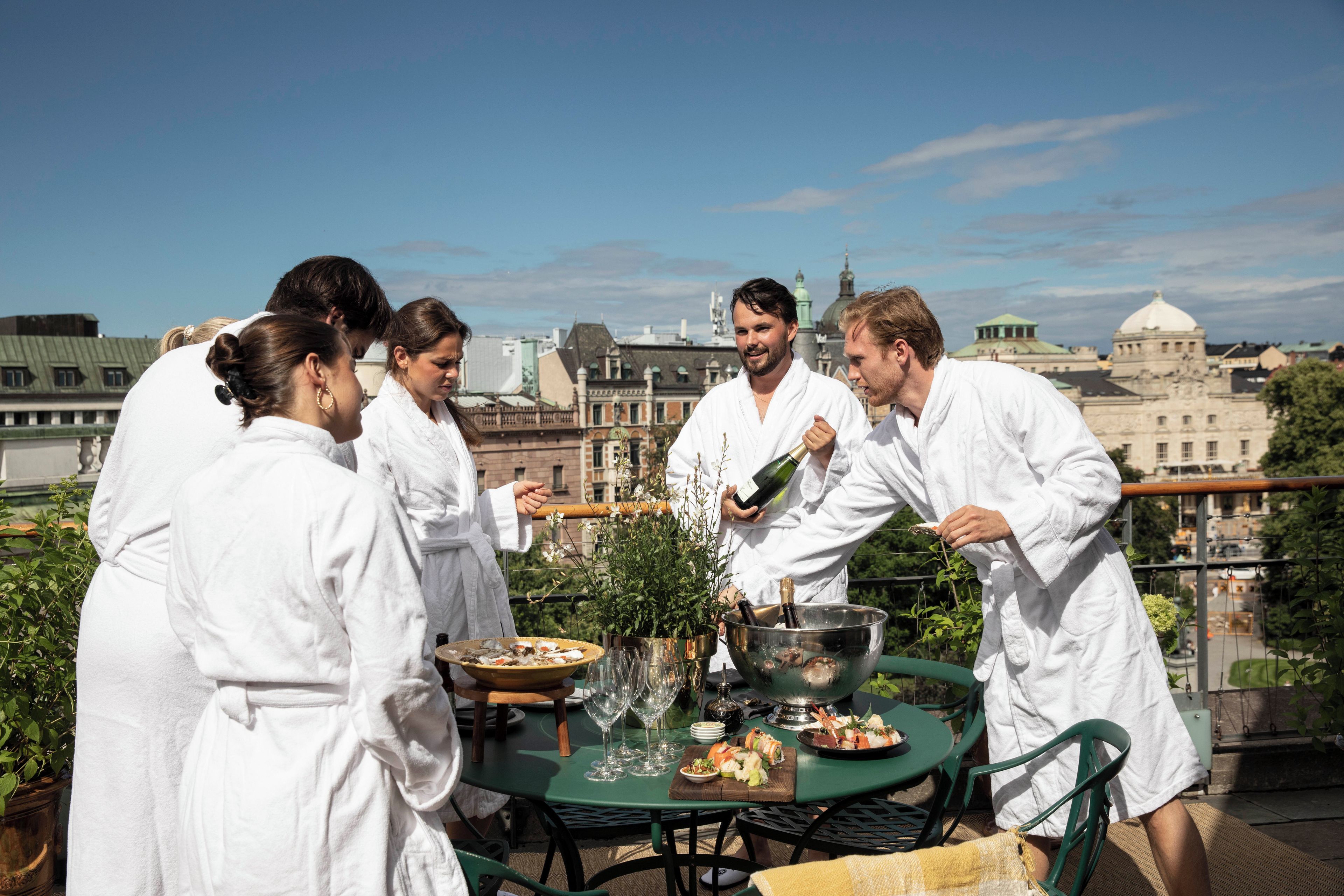A group of people in white robes gathered around a table with food and drinks on a rooftop terrace with a cityscape in the background.