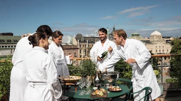 A group of people in white robes gathered around a table with food and drinks on a rooftop terrace with a cityscape in the background.