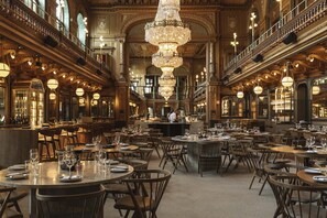 Un comedor espacioso y ornamentado con múltiples mesas y sillas, una lámpara de araña imponente y un balcón con vista al interior.