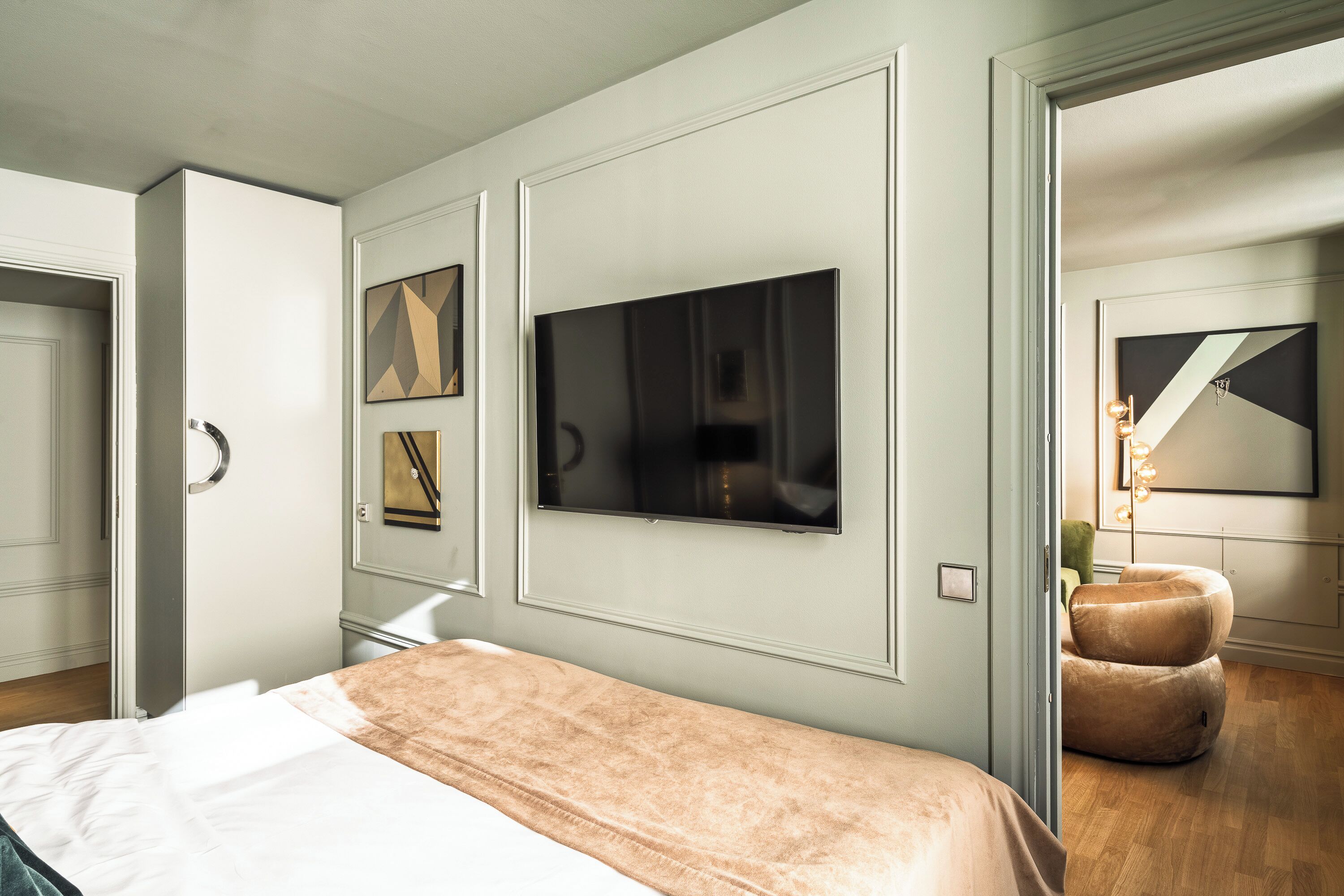 A modern bedroom with a large bed, a flat-screen TV mounted on the wall, and a seating area with a cushioned chair.