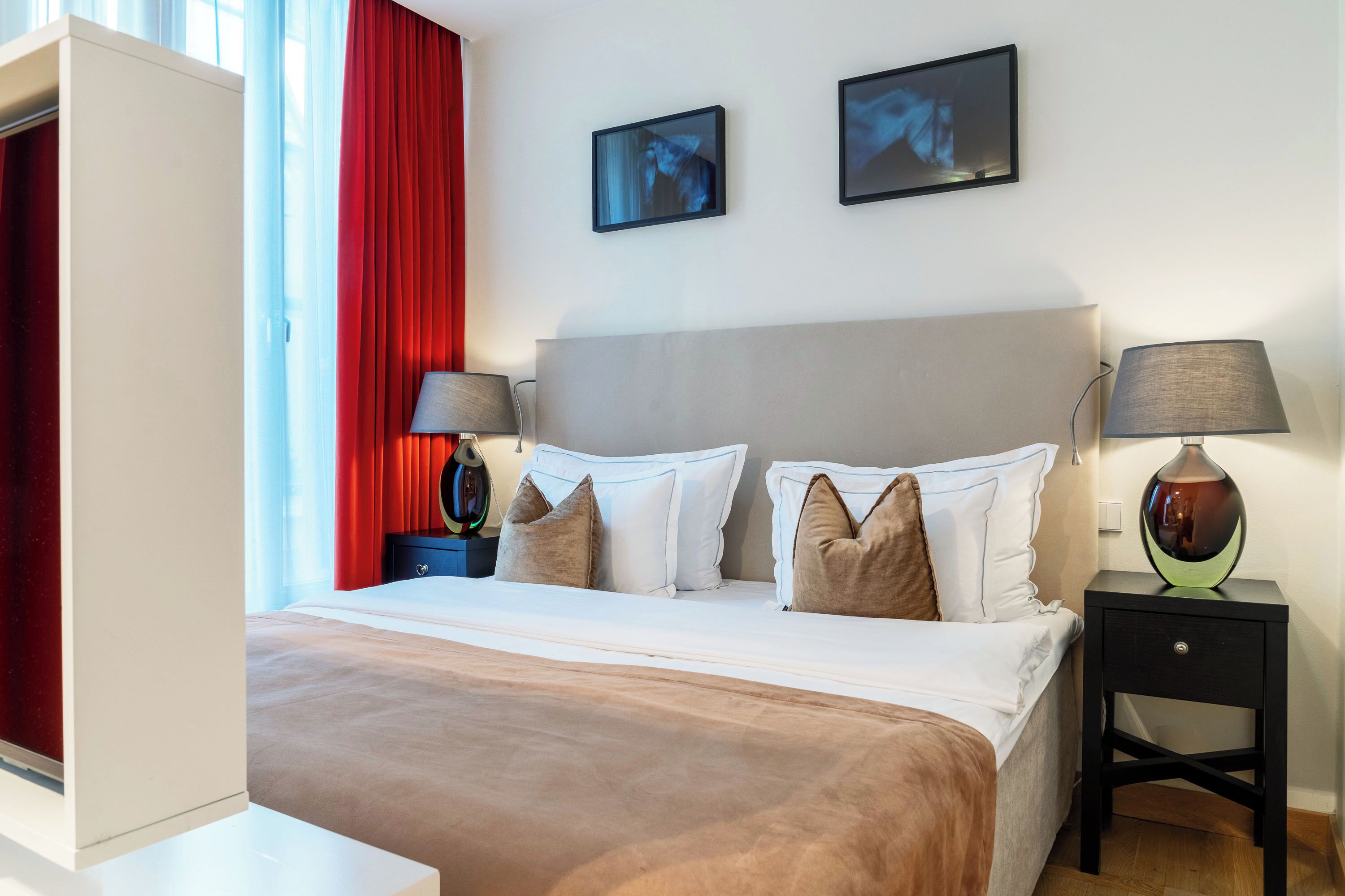 A hotel room with a bed, two bedside tables, a wardrobe, and two wall-mounted pictures.