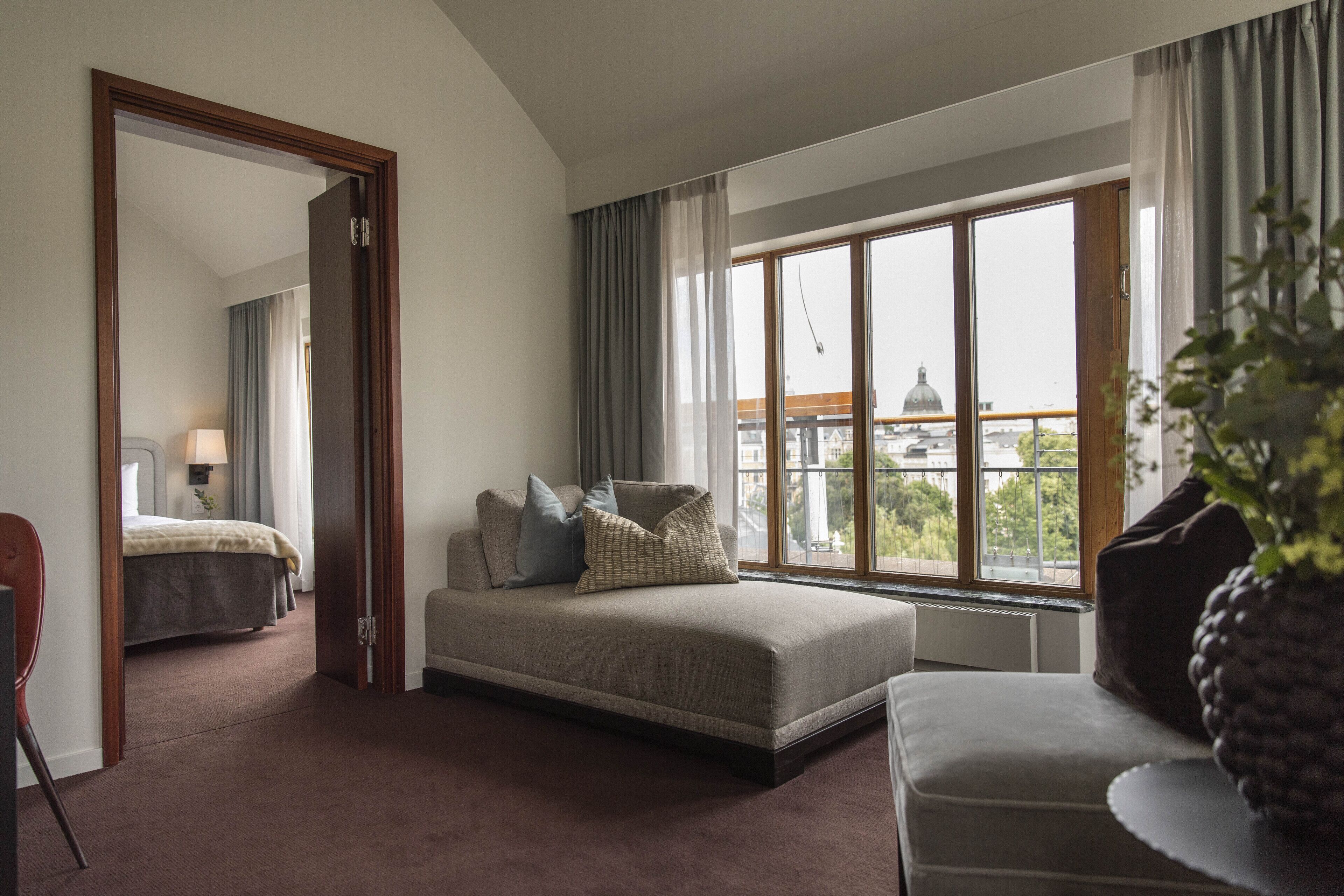 A modern hotel room with a large bed, a sofa, and a view of a cityscape through large windows.