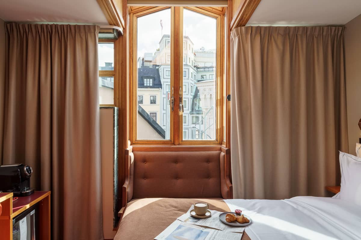 A hotel room with a bed, a coffee table with a cup and croissants, a newspaper, and a view of city buildings through a large window.