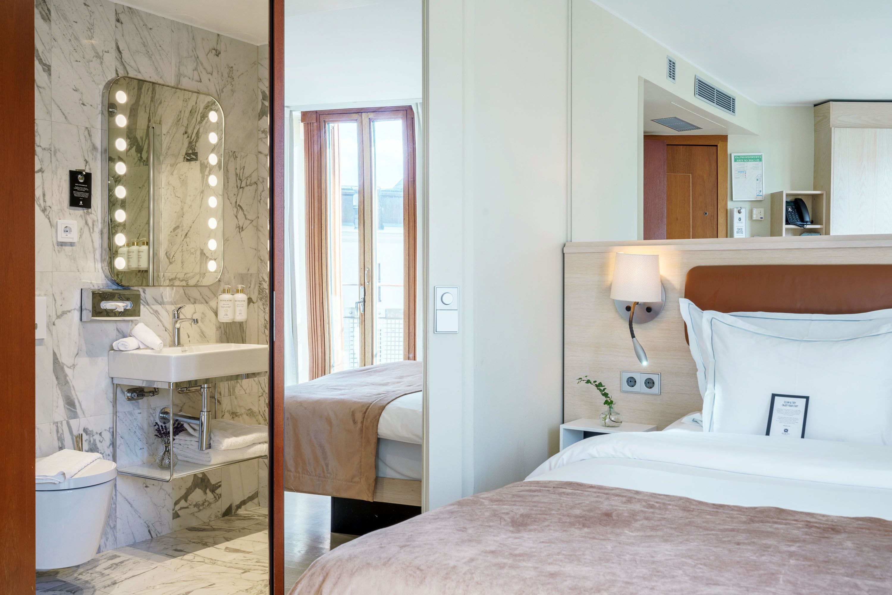 A hotel room with a marble bathroom, a bed with a headboard, a nightstand with a lamp, and a mirror with lights.