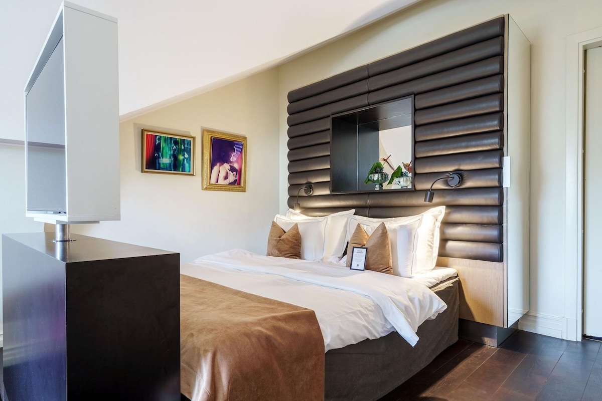 A modern hotel room with a large bed, a flat-screen TV, a wall-mounted TV, and two framed artworks.