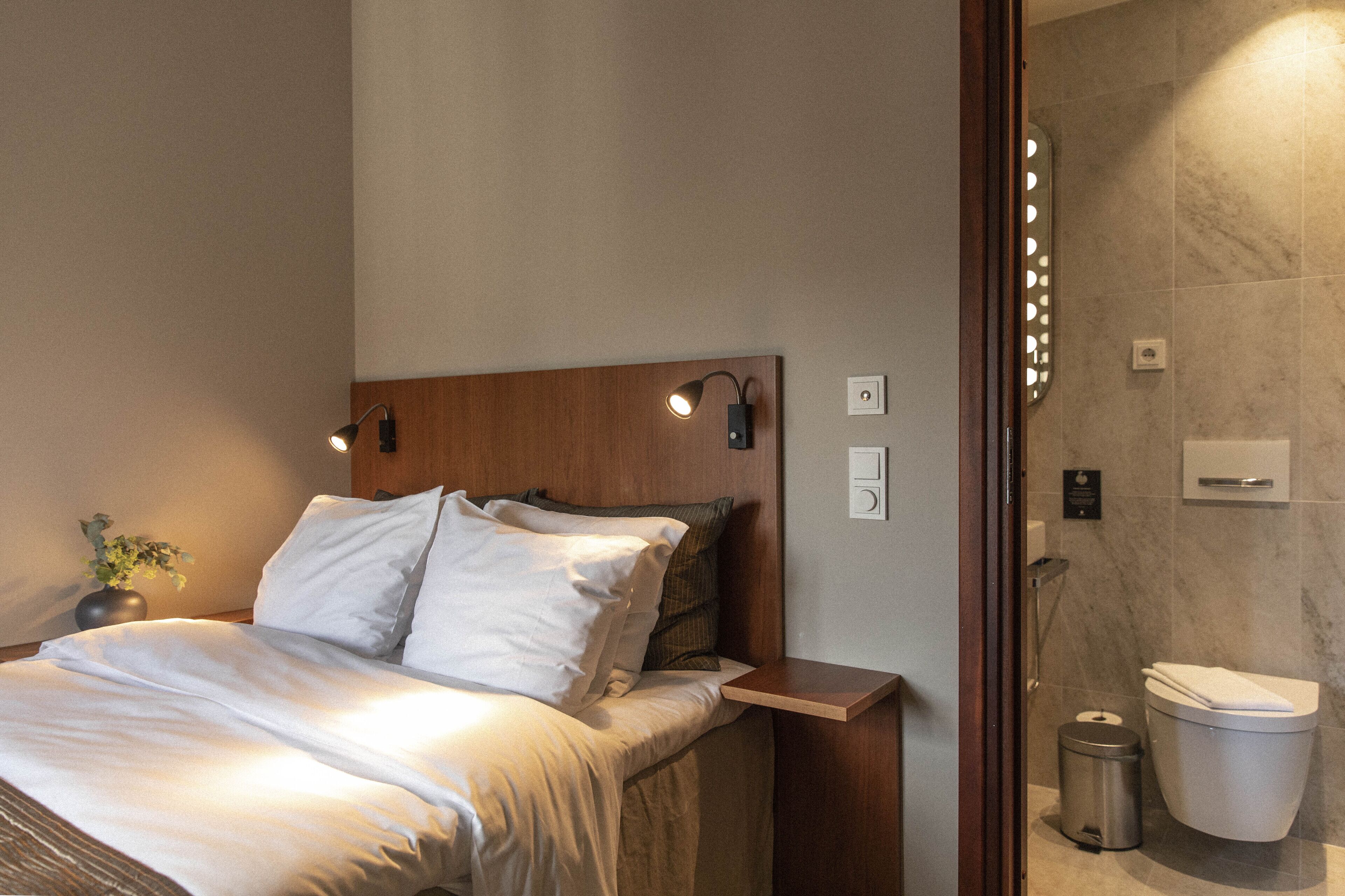A hotel room with a wooden headboard, a bedside table, a small trash bin, and a toilet.